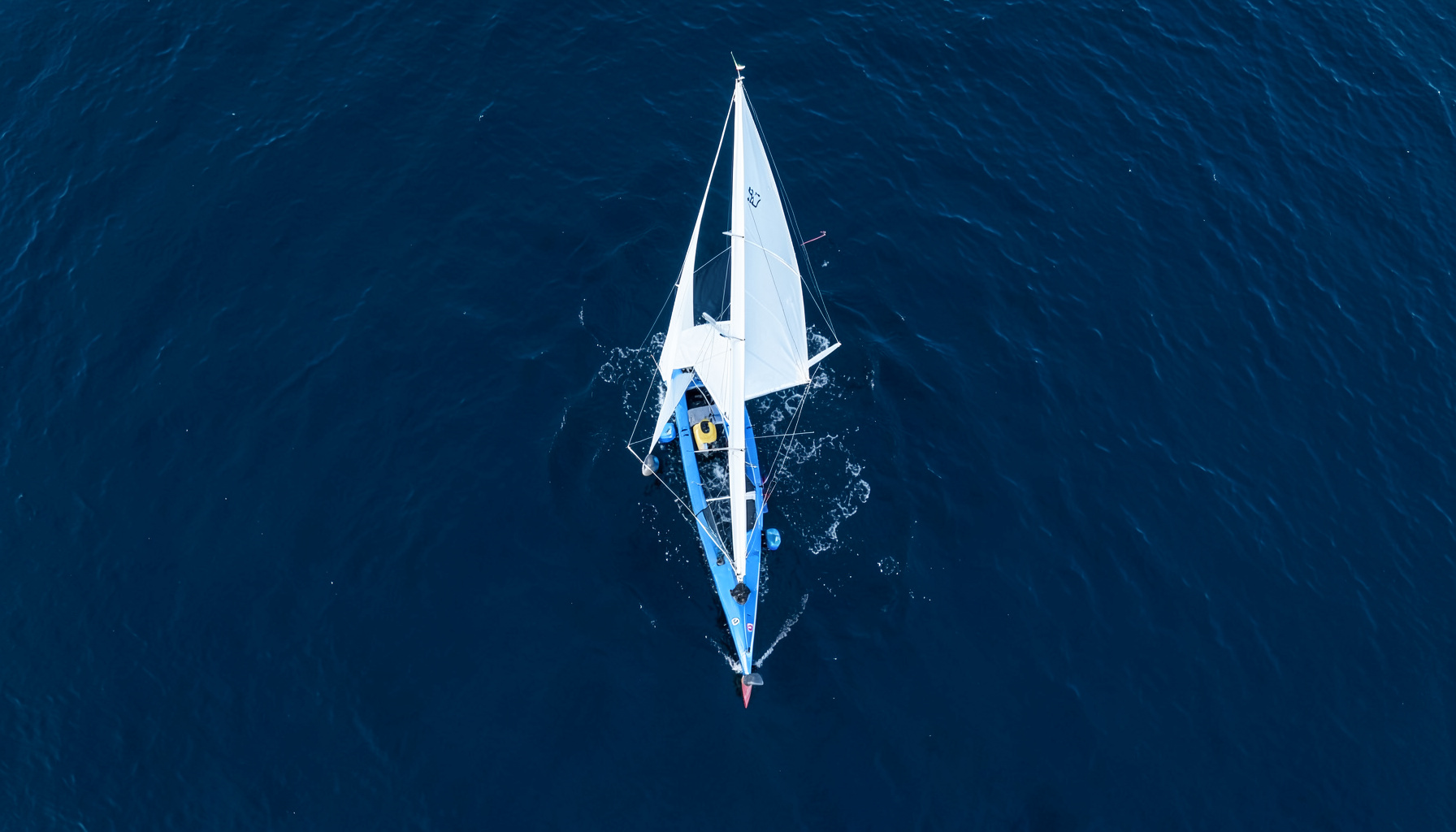Barca a vela Laser con vela bianca che taglia l'acqua blu scuro, vista zenitale dall'alto