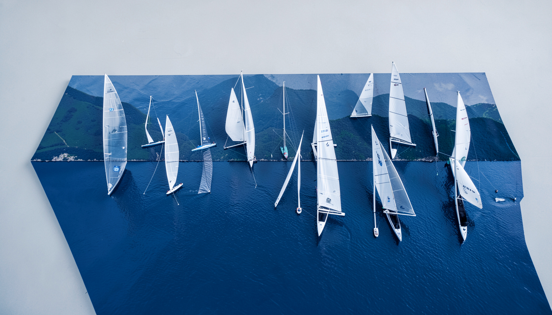 Regata di Laser sul lago con numerose vele bianche allineate, montagne verdi sullo sfondo