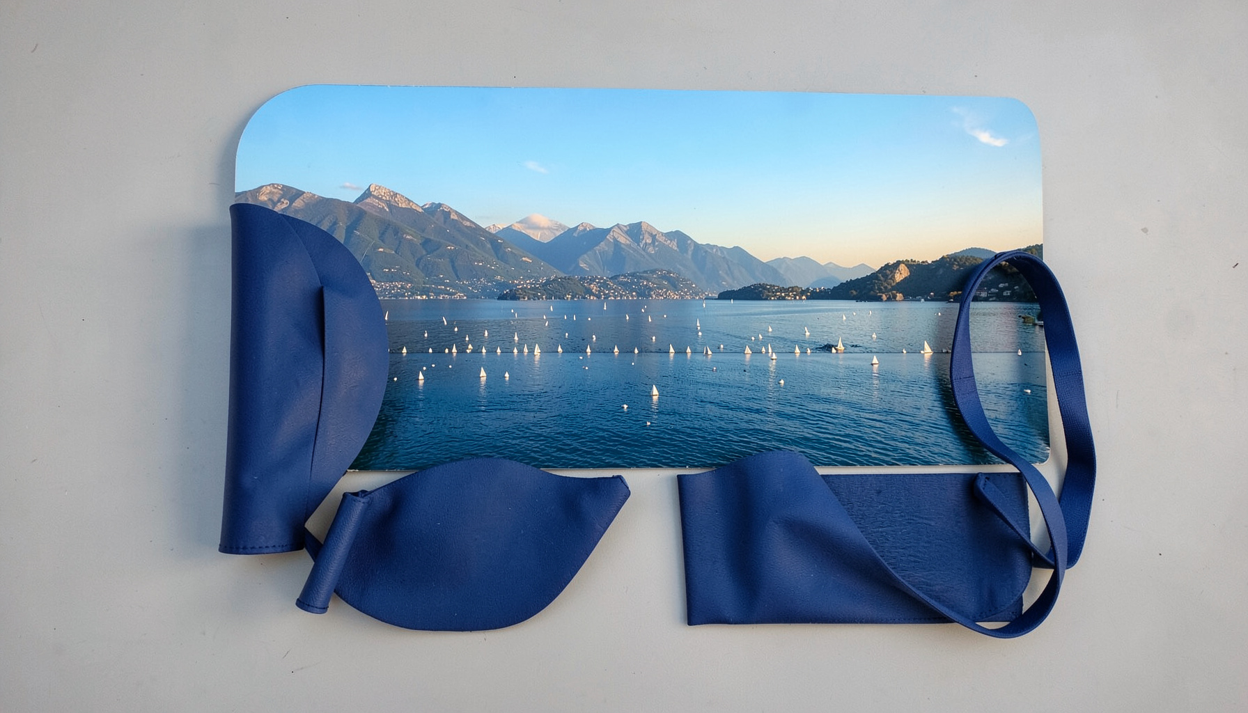 Vista panoramica del Lago di Garda con vele bianche in lontananza, cielo azzurro e montagne sullo sfondo, luce calda del sole